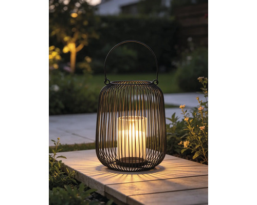 Modernes Windlicht aus schwarzem Metalldraht mit Glaseinsatz und leuchtender Kerze auf einer Gartenplatte.