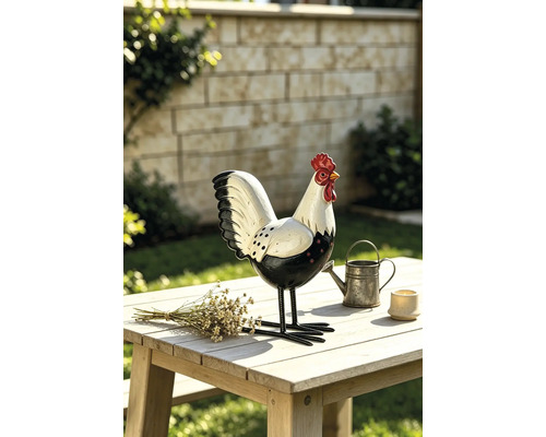 Dekorative Gartenfigur Hahn aus Metall auf einem hellen Holztisch im Garten neben einer kleinen Gießkanne und Trockenblumen.