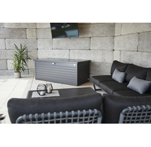 Metall-Auflagenbox auf einer hellen Terrasse mit Loungesofa, Beistelltisch, einer Pflanze und einer Wand aus Steinblöcken.