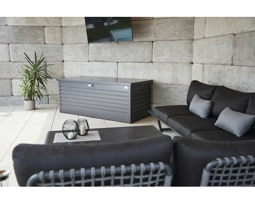 Metall-Auflagenbox auf einer hellen Terrasse mit Loungesofa, Beistelltisch, einer Pflanze und einer Wand aus Steinblöcken.