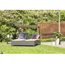 Doppel-Sonnenliege aus Polyrattan mit Sonnendach und Kissen in einem hellen Garten mit Rasen, blühenden Sträuchern und Brunnenwand aus Metall.