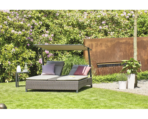 Doppel-Sonnenliege aus Polyrattan mit Sonnendach und Kissen in einem hellen Garten mit Rasen, blühenden Sträuchern und Brunnenwand aus Metall.