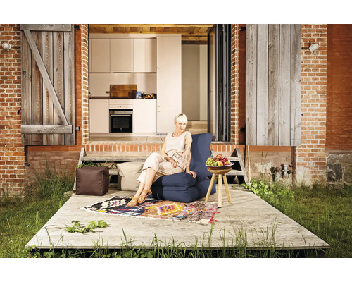 Frau sitzt auf einem blauen Sitzsack auf einer Holzterrasse vor einem Haus mit Blick in die Küche, neben Sitzwürfeln und einem bunten Teppich.