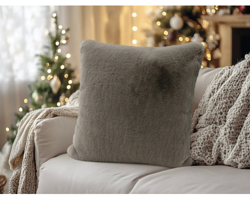 Weiches, graues Kunstfell-Kissen auf einem Sofa in weihnachtlicher Atmosphäre mit einem geschmückten Tannenbaum im Hintergrund.