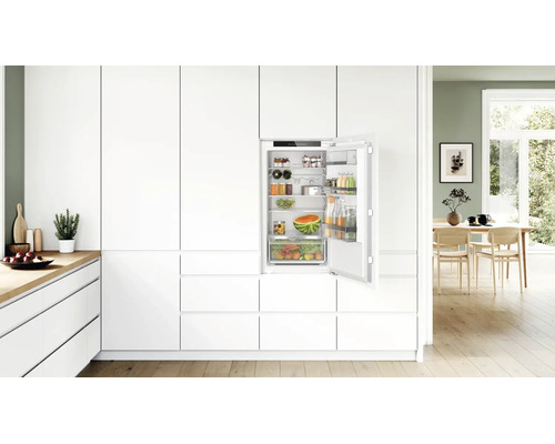 Einbaukühlschrank in moderner, weißer Einbauküche mit offener Tür und gefüllten Regalfächern und Gemüsefach.