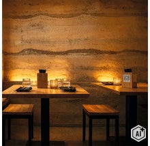 Restaurant mit Holztischen und Stühlen vor einer strukturierten Stampflehmwand mit atmosphärischer indirekter Beleuchtung. (Image AI modified or generated)