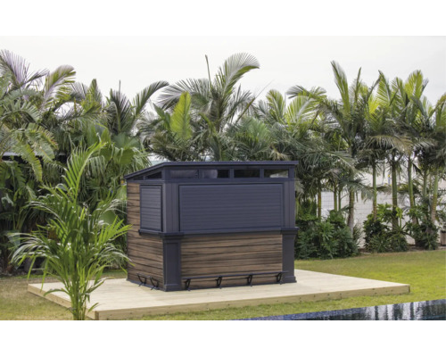 Moderne Gartenbar mit Holzoptik und dunklen Rahmenelementen auf einer Holzterrasse im Garten vor einer tropischen Bepflanzung.