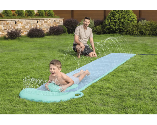 Ein Kind rutscht auf einer Wasserrutsche mit Sprühfunktion über eine Rasenfläche im Garten, während ein Mann im Hintergrund zuschaut.