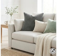 Ein helles Sofa mit Dekokissen in Cordstruktur und Stoffoptik sowie einer Strickdecke in einem Wohnzimmer. (Image AI modified or generated)