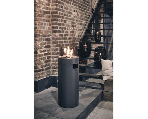 Runder Gas-Terrassenkamin mit Glaseinsatz auf einer Terrasse vor einer Backsteinwand mit Treppe und dekorativen Laternen.