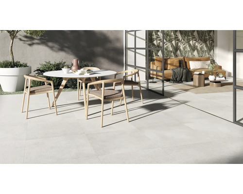 Moderne Terrasse mit hellen Bodenfliesen, runder Tischgruppe und Übergang in einen lichtdurchfluteten Wohnbereich mit raumhoher Verglasung.
