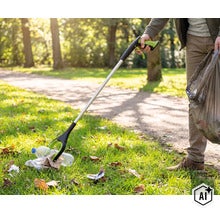 Person verwendet eine Müllgreifhilfe im Park, um eine Plastikflasche vom Boden aufzuheben und in einen Müllbeutel zu legen. (Image AI modified or generated)