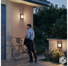 Ein Mann mit Fahrrad vor einem Haus bei Abenddämmerung, beleuchtet durch zwei moderne quadratische Außenwandleuchten an der Fassade. (Image AI modified or generated)