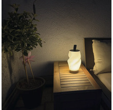 Moderne LED Tischleuchte mit gewelltem Lampenschirm auf einem Holztisch neben einer Topfpflanze und Gartenmöbeln bei Nacht