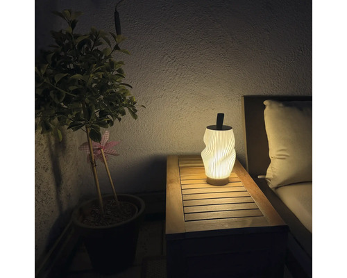 Moderne LED Tischleuchte mit gewelltem Lampenschirm auf einem Holztisch neben einer Topfpflanze und Gartenmöbeln bei Nacht