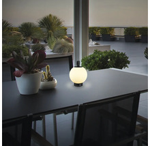 Moderne runde LED Tischleuchte für den Außenbereich auf einem Terrassentisch neben einer Pflanze bei Dämmerung.