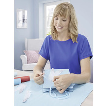 Eine Frau beim Verpacken eines Geschenks mit einer Geschenktüte und dekorativen Federn in einem hell eingerichteten Raum.