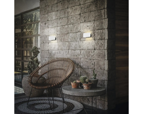 Zwei moderne LED Außenwandleuchten montiert an einer Steinwand über einem gemütlichen Lounge Bereich mit Rattansessel und Beistelltisch.