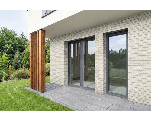 Moderne Hausfassade mit Klinkerriemchen, bodentiefen Fenstern und einer Holzstütze auf einer Terrasse vor einer Gartenlandschaft.