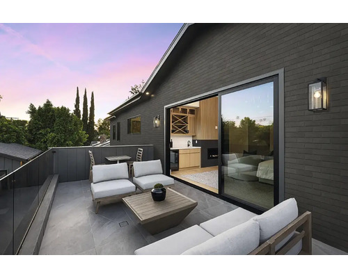 Moderne Dachterrasse mit Lounge Mobiliar, Steinfliesen und Schiebetür zum Wohnbereich in einem Haus mit dunkler Ziegelfassade bei Dämmerung.