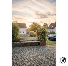 Eine Gartenbank aus Holz vor einer Steinmauer neben einem hohen Pflanzgefäß mit Rankgitter und blühenden Pflanzen auf einem gepflasterten Hof. (Image AI modified or generated)