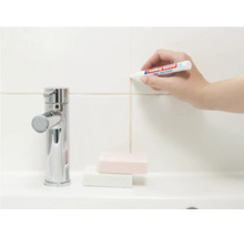 Eine Hand verwendet einen Edding Fugenmarker zur Auffrischung der Fliesenfugen an einer Wand neben einem Wasserhahn.