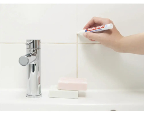 Eine Hand verwendet einen Edding Fugenmarker zur Auffrischung der Fliesenfugen an einer Wand neben einem Wasserhahn.