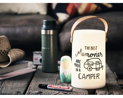 Eine dekorative Laterne mit Tragegriff und dem Schriftzug The Best Memories Are Made In A Camper auf einem Holztisch mit Stift.