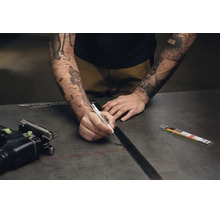 Eine Person zeichnet mit einem Edding Tieflochmarker eine Linie an einem Metalllineal auf einer dunklen Arbeitsplatte.