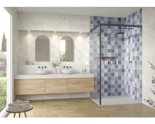 Helles Badezimmer mit Waschtisch, zwei Aufsatzwaschbecken, zwei Spiegeln und einer Duschkabine mit blau-weiß gemusterten Fliesen.