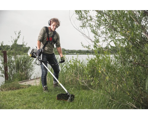 Mann verwendet einen Rasentrimmer mit Tragegurt und Schutzausrüstung in einem Garten am See