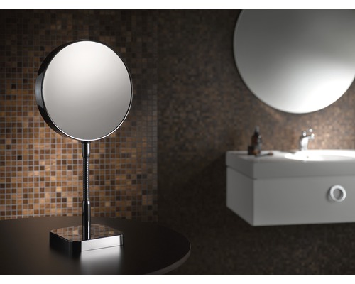 Runder Kosmetikspiegel auf einem Tisch, mit Waschbecken und Spiegel im Hintergrund.