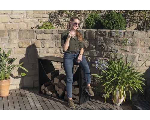 Frau sitzt auf einem Brennholzregal im Freien mit Mauerwerk im Hintergrund.