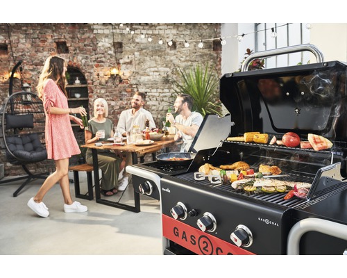 Szene mit einem Gasgrill, auf dem verschiedene Speisen zubereitet werden, während eine Gruppe von Menschen im Hintergrund an einem Tisch sitzt.