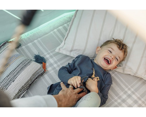 Lachender Junge liegt auf einer Decke mit Kissen und spielt mit Dinosaurier Spielzeug