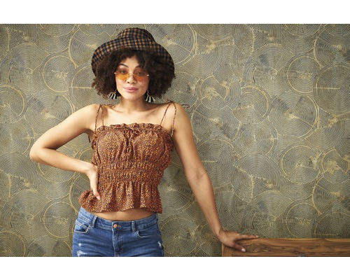 Frau mit Hut, gemustertem Oberteil und Jeans vor einer gemusterten Tapete