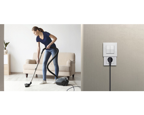 Frau saugt mit einem Staubsauger einen Teppich in einem hellen Wohnzimmer. Eine Steckdose und ein Lichtschalter sind an der Wand befestigt.