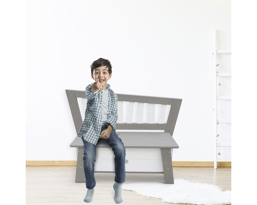 Junge sitzt auf einer grauen Spielzeugtruhenbank und zeigt mit dem Finger.