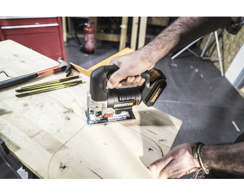 Eine Person sägt mit einer Akku-Stichsäge von Worx ein Holzbrett. Werkzeuge wie Hammer und Zollstock liegen bereit.
