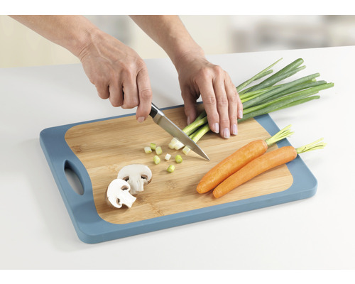 Hände schneiden Frühlingszwiebeln auf einem Schneidebrett mit Messer, neben Karotten und Champignons.