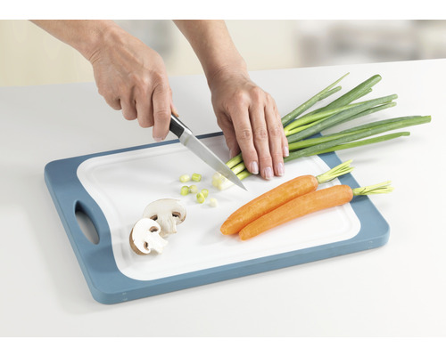 Eine Person schneidet auf einem Schneidebrett Lauchzwiebeln mit einem Messer. Karotten und Champignons liegen ebenfalls auf dem Brett.