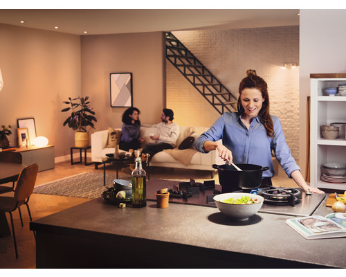 Helle, offene Wohnküche mit Frau, die am Herd kocht und Personen im Hintergrund auf dem Sofa sitzend.