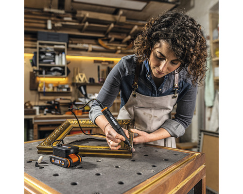Frau arbeitet mit einem Akku-Multifunktionswerkzeug von Worx an einem goldenen Bilderrahmen in einer Werkstatt.