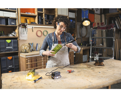 Frau bohrt mit einem Multifunktionswerkzeug ein Loch in eine Glasflasche in einer Werkstatt.