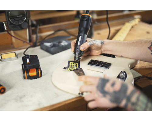 Ein Mann entfernt mit einem Heißluftgebläse einen Aufkleber von einer Gitarre. Ein Werkzeug zum präzisen Erhitzen befindet sich auf einer Werkbank.