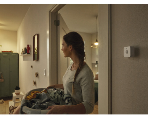 Eine Frau trägt einen Wäschekorb durch einen Flur, an der Wand ist ein Smart Home Bewegungsmelder montiert.