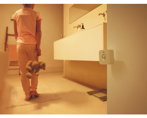 Ein weißer quadratischer Bewegungsmelder an der Wand in einem schwach beleuchteten Badezimmer mit einer Person, die einen Teddybären hält.