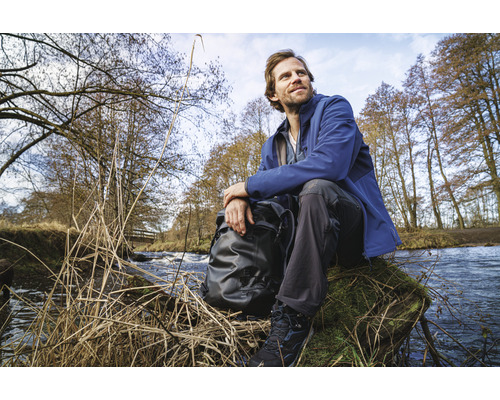 Ein Mann sitzt mit Rucksack an einem Bachufer in der Natur.