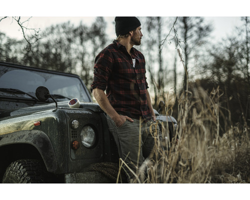 Ein Mann in kariertem Hemd lehnt an einem Geländewagen in einer ländlichen Umgebung.