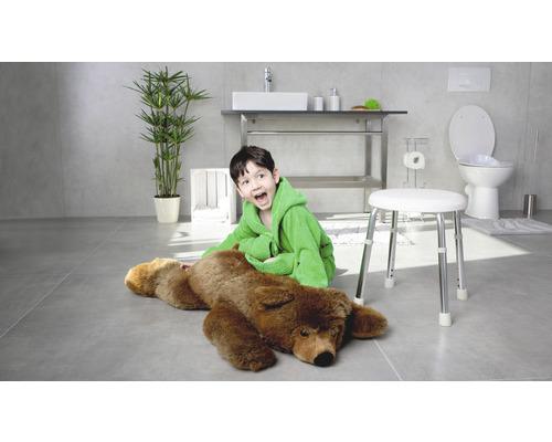 Helles Badezimmer mit einem Jungen im Bademantel, der neben einem Bären auf einem Hocker sitzt, mit Waschbecken, Toilette und Pflanze